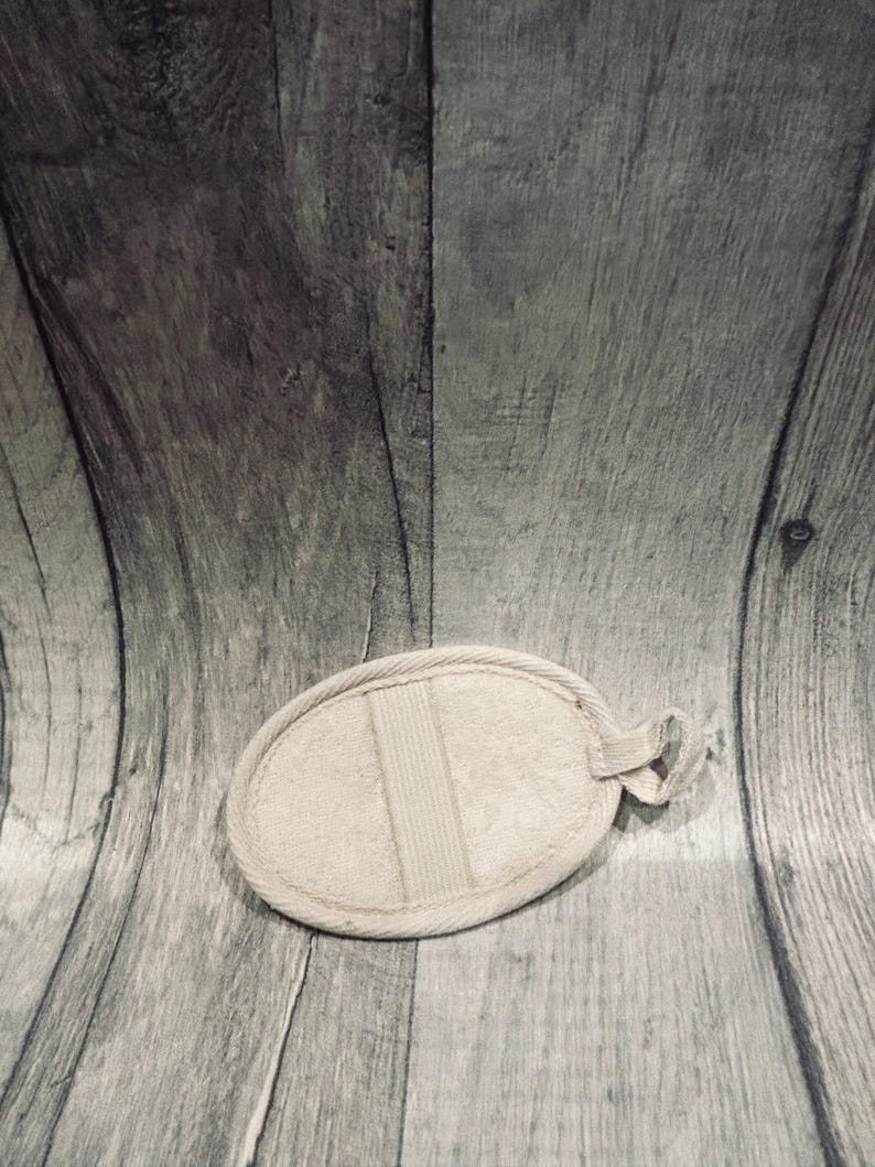 May include: An oval-shaped, beige bath sponge with a loop for hanging. The sponge has a textured surface and a vertical band across the center. The background is a gray wooden surface.
