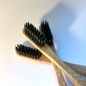 May include: Close-up of three bamboo toothbrushes with black bristles. The toothbrushes are made of light brown bamboo and have a simple, minimalist design. The bristles are densely packed and appear soft. The toothbrushes are arranged in a slightly overlapping pattern.