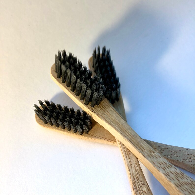 May include: Close-up of three bamboo toothbrushes with black bristles. The toothbrushes are made of light brown bamboo and have a simple, minimalist design. The bristles are densely packed and appear soft. The toothbrushes are arranged in a slightly overlapping pattern.