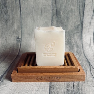 Op de afbeelding: Een witte zeep in een houten zeepbakje. De zeep heeft een textuur en is bedrukt met een logo en tekst. Het houten bakje heeft een lattenontwerp en de tekst "Ellie's Eco Shop Zero Waste Living".