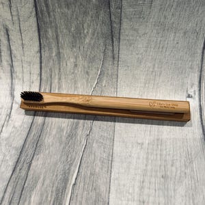 May include: A bamboo toothbrush with black bristles rests in a cardboard holder. The toothbrush handle is light brown, and the holder is a similar shade. The text "Ellie's Eco Shop Zero Waste Living" is printed on the holder.
