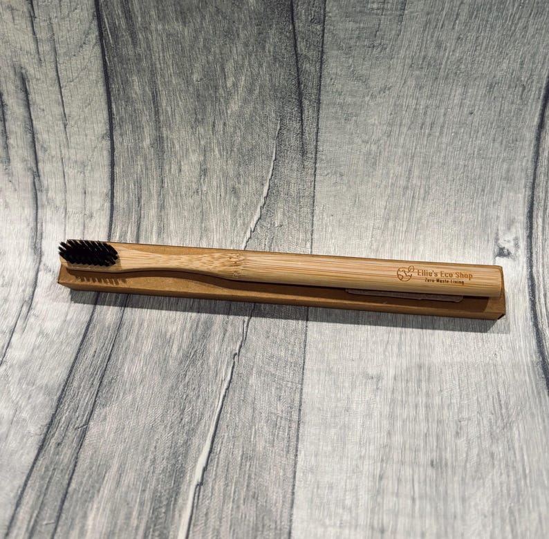 May include: A bamboo toothbrush with black bristles rests in a cardboard holder. The toothbrush handle is light brown, and the holder is a similar shade. The text "Ellie's Eco Shop Zero Waste Living" is printed on the holder.