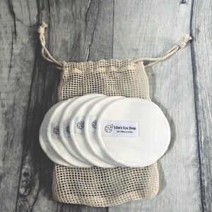 May include: A set of white, reusable facial rounds with the text "Ellie's Eco Shop" on a small label. The rounds are stacked and placed inside a beige mesh bag with a drawstring closure. The background is a gray wooden surface.