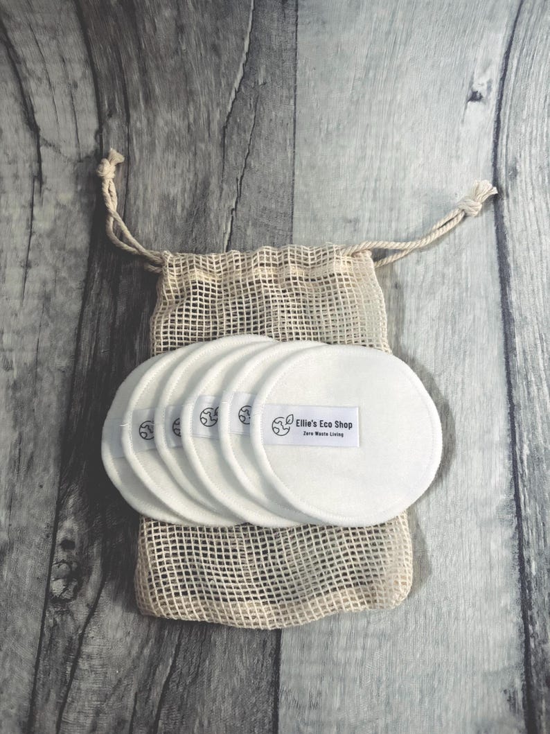 May include: A set of white, reusable facial rounds with the text "Ellie's Eco Shop" on a small label. The rounds are stacked and placed inside a beige mesh bag with a drawstring closure. The background is a gray wooden surface.