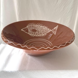 Könnte beinhalten: Eine Terrakotta-Schüssel mit einem weißen Fischdesign. Die Schüssel hat einen gewellten Rand und ein weißes Kreisdesign in der Mitte.