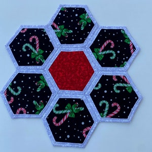 May include: A Christmas-themed table runner made of fabric with a hexagonal design. The runner is made of six black fabric hexagons with white outlines, each featuring a pattern of candy canes and green bows on a black background with white dots. A red hexagon with a white outline is in the center.
