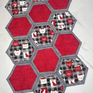 May include: A handmade, quilted table runner composed of hexagon-shaped fabric pieces. The design features alternating red and patterned panels. The patterned panels display a black and white plaid with cat and coffee cup motifs. The edges are finished with gray binding.