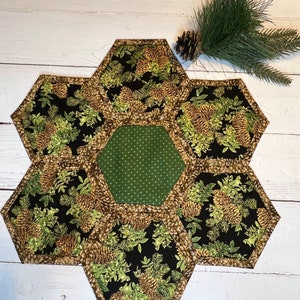 May include: A hexagonal table runner made of fabric with a pine cone pattern. The runner is made of six hexagonal pieces of fabric, each with a different pine cone design. The center piece is a solid green fabric with white polka dots.