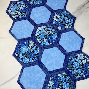 May include: A handmade, quilted table runner featuring a honeycomb design. The runner is composed of blue and floral patterned hexagons. The floral patterns include white and light blue flowers on a dark blue background. The edges are trimmed with a dark blue denim-like fabric.