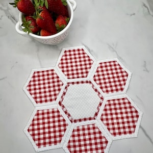 May include: Red and white gingham fabric hexagon table runner with white trim. The runner is made of six hexagons, with a white hexagon in the center.