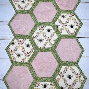 May include: A hexagonal table runner with a bee and floral pattern. The runner is made of fabric with a pink, green, and white color scheme. The runner is made up of individual hexagonal pieces sewn together.