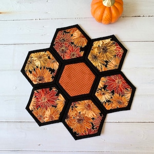 May include: A handmade, floral-patterned trivet with a black border, featuring a central hexagon with a dotted design. The trivet is surrounded by six hexagons with a floral pattern in shades of orange, yellow, and red. An orange pumpkin is in the background.
