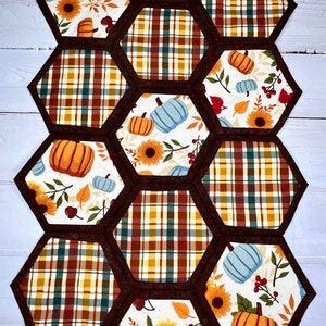 May include: A brown and white table runner with a hexagonal pattern. Each hexagon features a different autumn-themed design, including pumpkins, sunflowers, and plaid fabric.