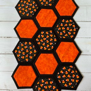 May include: A Halloween table runner made of fabric with a honeycomb pattern. The runner is made of orange and black fabric with a candy corn print. The runner is approximately 18 inches long and 12 inches wide.
