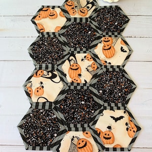 May include: A Halloween-themed table runner with a black and white checkered pattern and a black background with white glitter. The runner is made up of hexagonal fabric pieces with different designs, including pumpkins, bats, and spiderwebs.