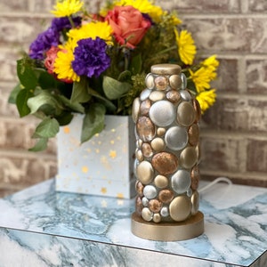 Op de afbeelding: Een tafellamp van goud, zilver en koperkleurige ronde glazen kralen. De lamp staat op een marmeren tafelblad.