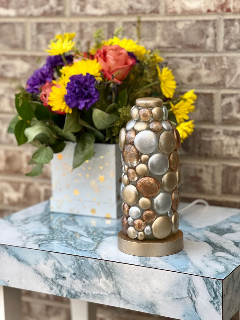 Op de afbeelding: Een tafellamp van goud, zilver en koperkleurige ronde glazen kralen. De lamp staat op een marmeren tafelblad.
