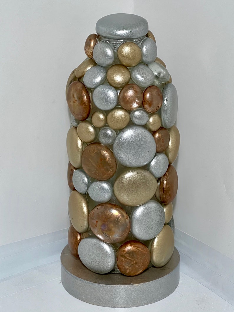 Op de afbeelding: Een decoratieve glazen fles bedekt met ronde stenen in zilver, goud en koperkleuren. De fles heeft een zilveren voet.
