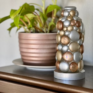 Op de afbeelding: Een decoratieve vaas van zilver en goud gemaakt van ronde stenen. De vaas staat op een houten tafel met een plant in een pot op de achtergrond.