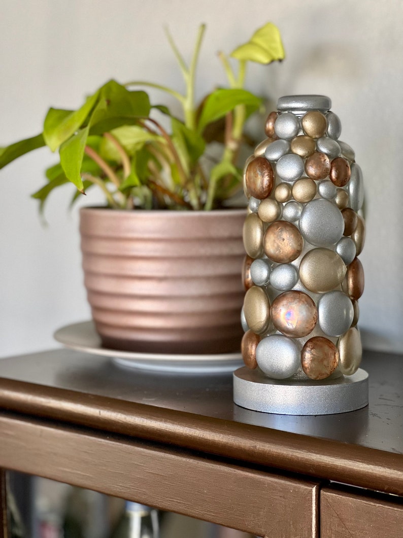 Op de afbeelding: Een decoratieve vaas van zilver en goud gemaakt van ronde stenen. De vaas staat op een houten tafel met een plant in een pot op de achtergrond.