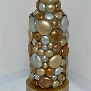 Op de afbeelding: Een decoratieve fles met een gouden basis en een cilindrisch lichaam bedekt met ronde, metalen stenen. De stenen zijn in tinten goud, zilver en koper.