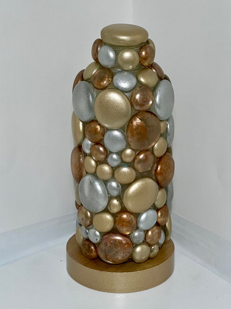 Op de afbeelding: Een decoratieve fles met een gouden basis en een cilindrisch lichaam bedekt met ronde, metalen stenen. De stenen zijn in tinten goud, zilver en koper.