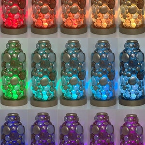 Op de afbeelding: Een set van 10 decoratieve glazen lampen met een uniek ontwerp. Elke lamp is gemaakt van een heldere glazen cilinder bedekt met kleine, ronde, iriserende glazen kralen. De lampen worden van binnenuit verlicht, waardoor een kleurrijk en betoverend effect ontstaat. De lampen zijn verkrijgbaar in verschillende kleuren, waaronder rood, oranje, geel, groen, blauw, paars en roze.