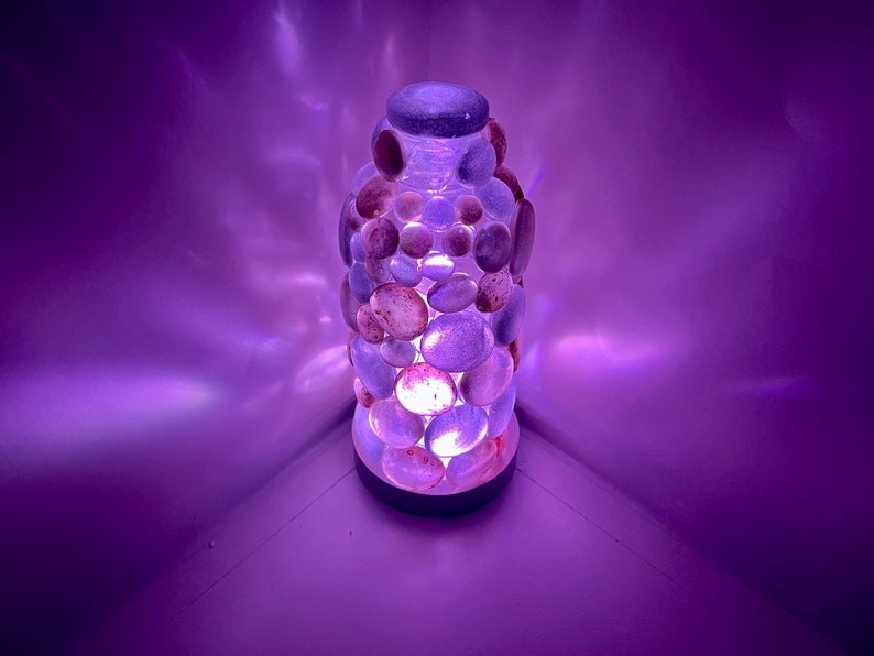 Op de afbeelding: Een paarse glazen lamp met een voet van kleine, gladde stenen. De lamp is aan en geeft een warm licht.