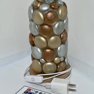 Op de afbeelding: Een tafellamp gemaakt van een glazen cilinder bedekt met ronde, gladde stenen in tinten goud, zilver en koper. De lamp heeft een houten voet en wordt geleverd met een afstandsbediening om de lichtkleur te veranderen.