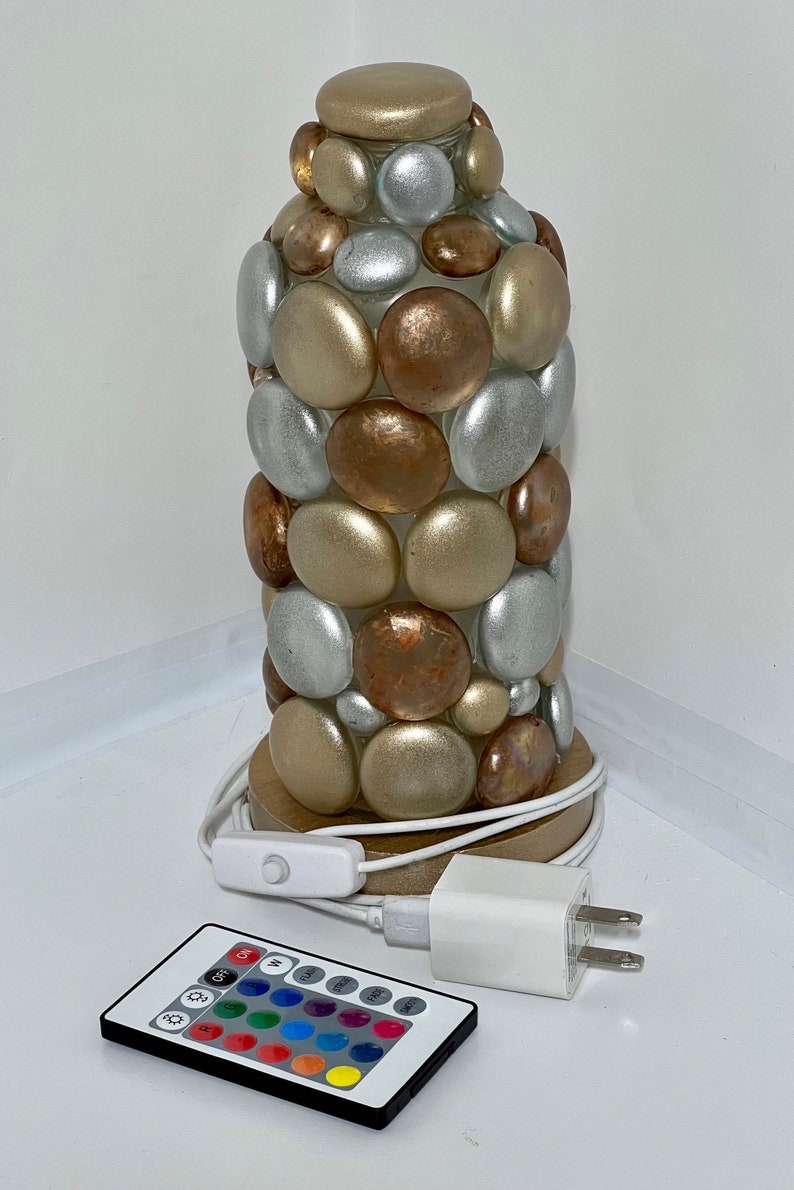 Op de afbeelding: Een tafellamp gemaakt van een glazen cilinder bedekt met ronde, gladde stenen in tinten goud, zilver en koper. De lamp heeft een houten voet en wordt geleverd met een afstandsbediening om de lichtkleur te veranderen.