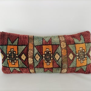 May include: A long, rectangular pillow with a geometric pattern in shades of red, orange, green, and brown. The pillow is made of a soft, woven fabric and has a fringed edge.