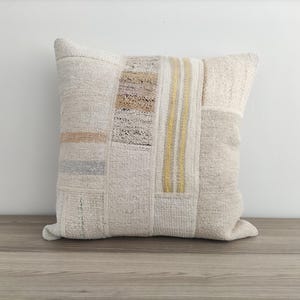 May include: A white throw pillow with a patchwork design featuring beige and yellow stripes. The pillow is made of a soft, textured fabric.