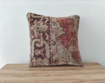 May include: Square cushion with a vintage rug design. The cushion features a faded red, beige, and burgundy colour palette. The cushion is placed on a wooden surface.