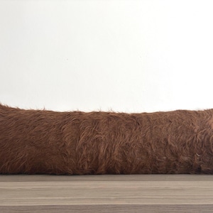 May include: A rectangular lumbar pillow featuring a long-pile, shaggy faux fur texture in a solid medium brown color. The pillow has a soft, dense appearance with fibers oriented in various directions across the entire surface of the cushion.