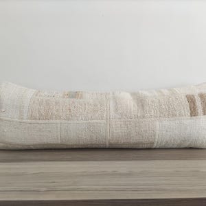 Op de afbeelding: Een lang, rechthoekig, wit kussen met een patchwork-ontwerp. Het kussen is gemaakt van verschillende tinten wit en beige stof en heeft een subtiele textuur.