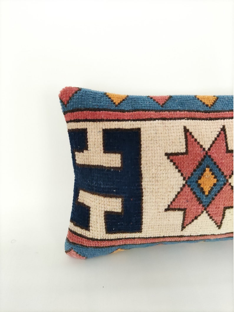May include: A long, rectangular pillow with a cream-colored background and a colorful geometric pattern. The pattern features blue, pink, and yellow colors in a repeating design.