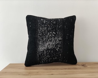 May include: Square black cushion with a central panel of white and black patterned fabric. The cushion is placed on a wooden surface against a white wall. The cushion is a decorative item.