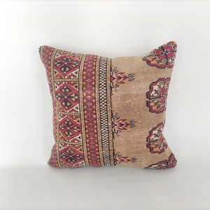 May include: A square pillow with a beige background and a red, black, and white geometric pattern. The pillow is made of a soft, woven fabric.