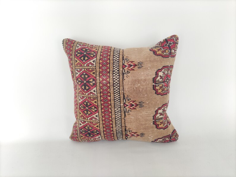 May include: A square pillow with a beige background and a red, black, and white geometric pattern. The pillow is made of a soft, woven fabric.