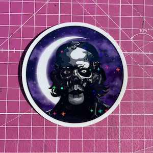 Puede incluir: Adhesivo redondo con un retrato estilizado en blanco y negro de una figura con apariencia de calavera, sobre un cielo estrellado púrpura y una luna creciente blanca. El adhesivo tiene un borde blanco y estrellas de colores.
