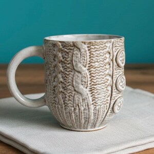 May include: A cream-colored ceramic mug with a textured cable knit design and button details. The mug has a curved handle and sits on a light gray napkin, with a teal background.
