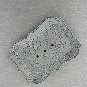 May include: A rectangular, light gray ceramic soap dish with scalloped edges and a raised inner platform. The surface is decorated with embossed patterns of circles, swirls, and sunbursts, and features three small drainage holes in the center.