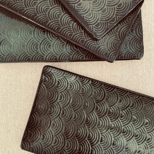 May include: A set of rectangular dark green ceramic plates featuring a repeating embossed wave pattern across the surface. The plates have a glossy finish and are arranged in a stack with one plate positioned separately in the foreground.
