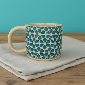 May include: A ceramic mug with a beige handle and rim, and a teal body decorated with a repeating floral pattern with yellow centers. The mug sits on a folded gray cloth, against a teal background.