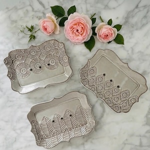May include: Three ceramic soap dishes with decorative floral and leaf patterns. The dishes are light gray with dark gray detailing and scalloped edges. Pink roses and green leaves are arranged around the dishes on a marble surface.