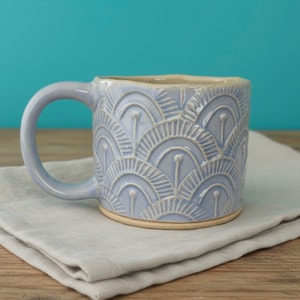 Puede incluir: Una taza de cerámica azul claro con asa, con un patrón repetitivo en forma de abanico. La taza está sobre una servilleta de lino gris, con un fondo azul turquesa. El borde y la base de la taza son beige.