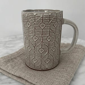 May include: A gray ceramic mug with a geometric pattern of diamonds and lines. The mug has a handle and sits on a tan linen napkin. The mug is approximately 4 inches tall.