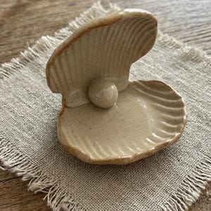 May include: A ceramic clam shell trinket dish with a glossy, speckled beige finish and a small, smooth pearl-like sphere resting in the center. The shell features a ribbed texture on both the top and bottom halves, with subtle brown edges.
