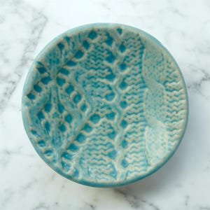 May include: A small, round, ceramic dish in a vibrant turquoise color. The dish features a textured, raised pattern resembling lace or a knitted design. The rim is slightly raised, and the overall aesthetic is handcrafted.
