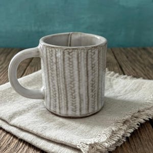 May include: A ceramic mug with a textured, vertical ribbed design in a light gray color. The mug has a rounded handle and sits on a folded, beige linen napkin. The background is a solid teal color, and the surface is a wooden table.